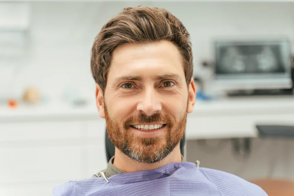 Bearded man with a relaxed smile sitting in a dental chair wearing a protective bib, with dental equipment and an X-ray screen blurred in the background - how much does it cost to have a tooth pulled
