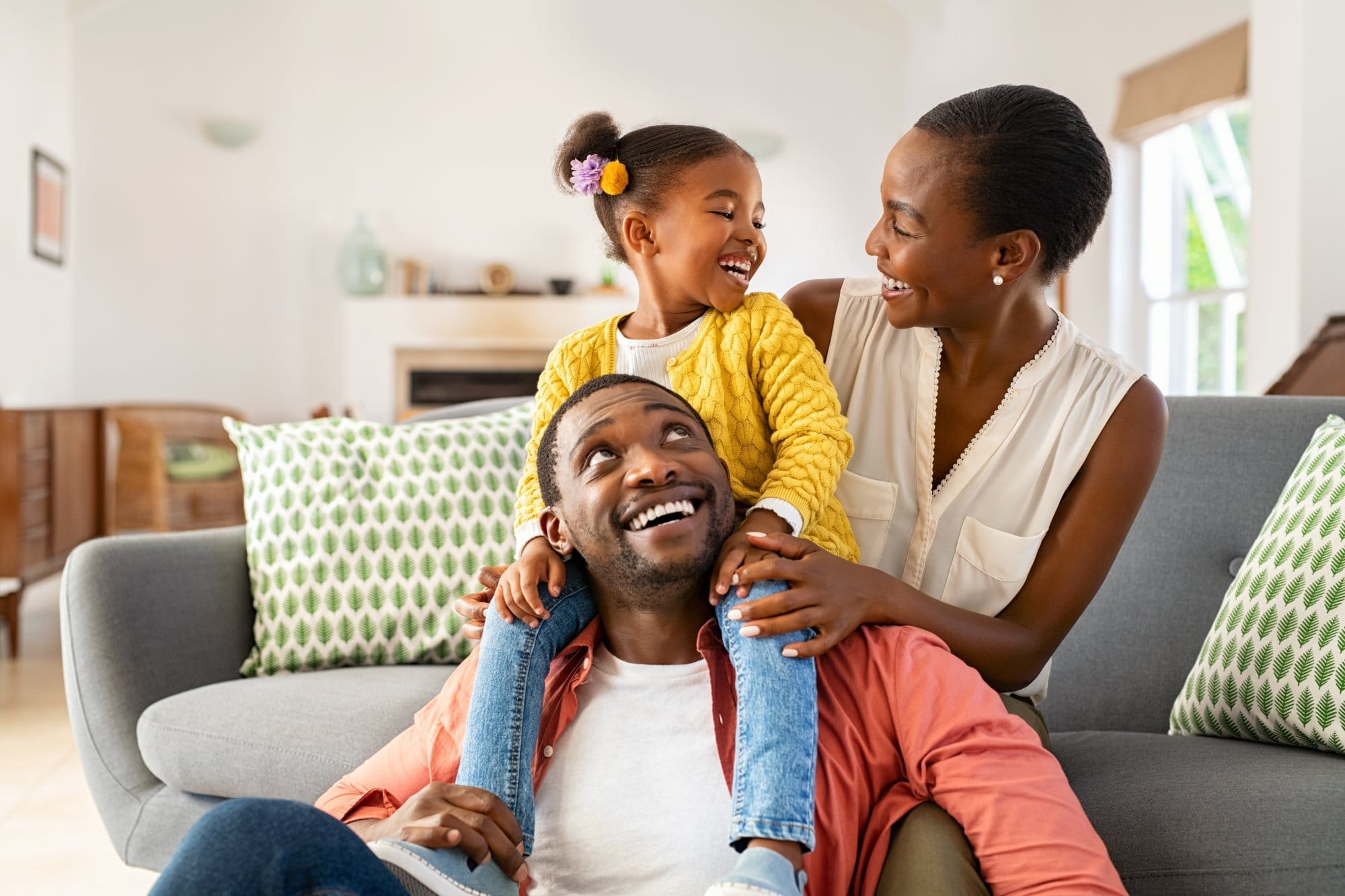 Cheerful little girl sitting on father shoulder while playing with mother at home. Happy black family enjoying weekend at home. Cute little daughter sitting on fathers shoulder and play with her mom.