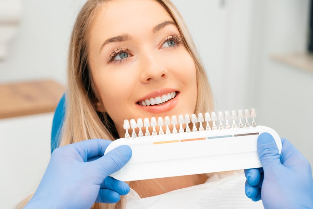 Blonde woman smiling in a dental chair while a gloved professional holds up a tooth shade guide to her smile for comparison during a veneer consultation - what do teeth look like under veneers