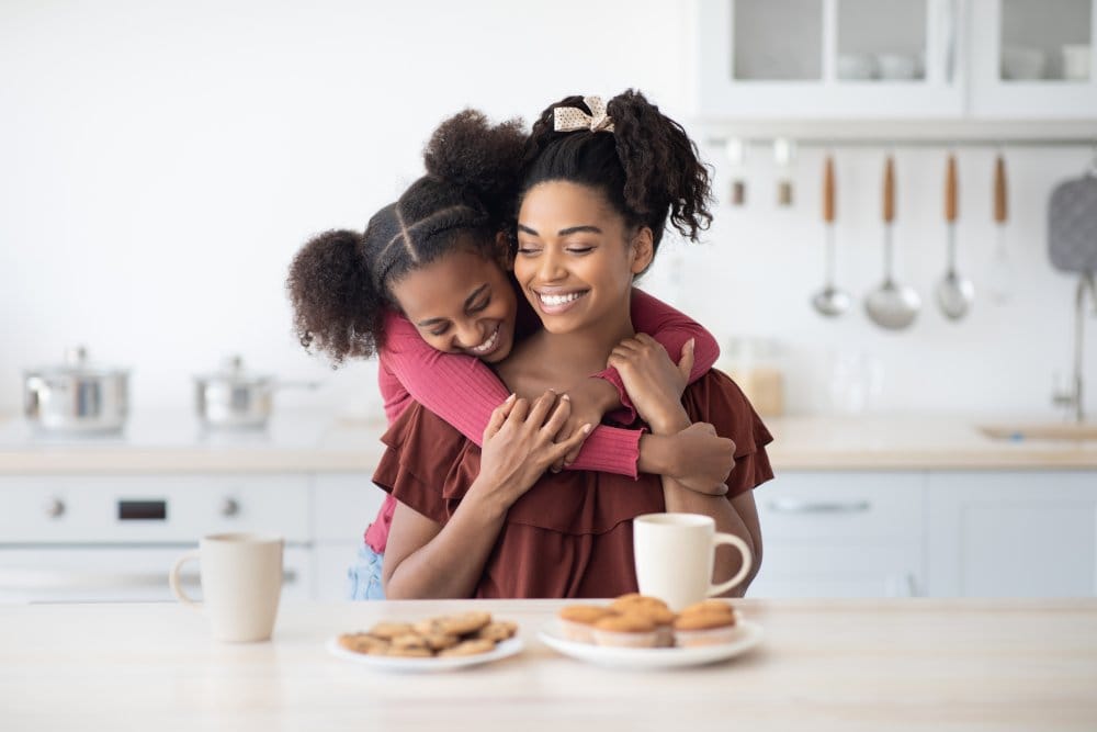 Emotional black daughter teenager hugging her beautiful happy mom while enjoying cookies, homemade pastry and biscuits together at home, kitchen interior. Family lifestyle, motherhood, parenthood