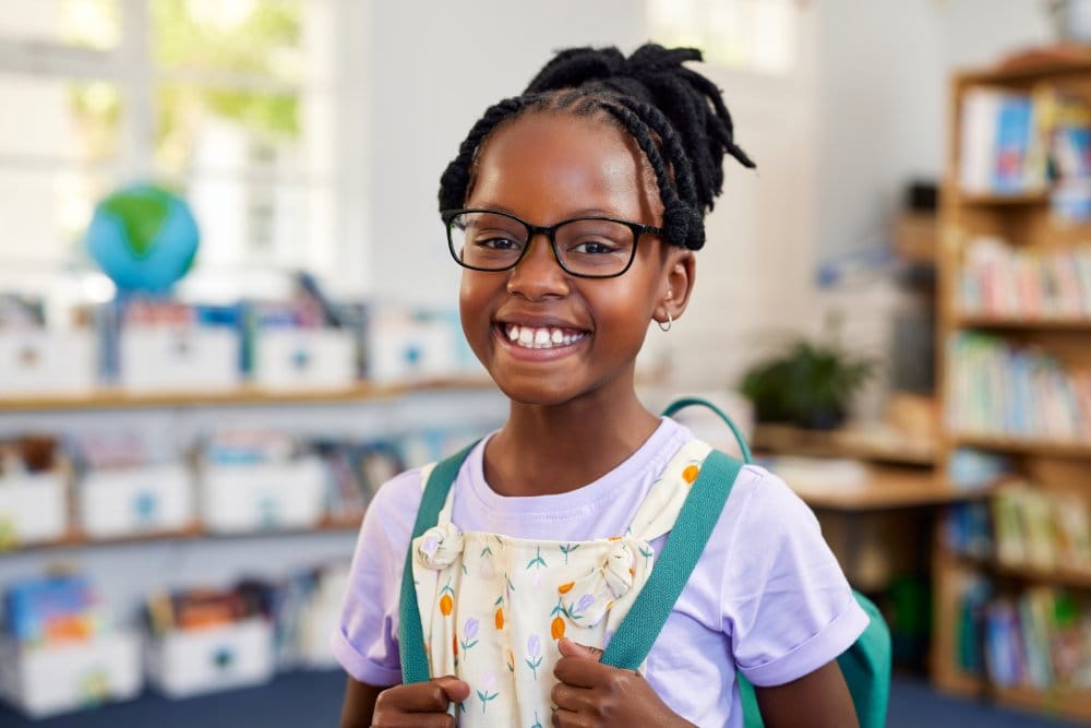 Portrait of a confident black girl wearing spectacles while holding backpack at library. Cheerful african american schoolgirl wearing eyeglasses standing in classroom while looking at camera.