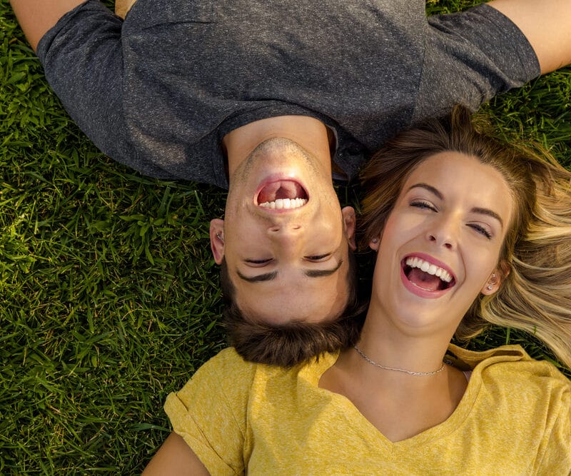 Happy young man and woman lying on green grass with heads close together, both laughing freely and enjoying a carefree moment - what does laughing gas do at the dentist