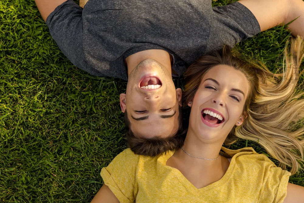 Young couple lying on grass laughing joyfully - what does laughing gas do at the dentist Happy young man and woman lying on green grass with heads close together, both laughing freely and enjoying a carefree moment - what does laughing gas do at the dentist