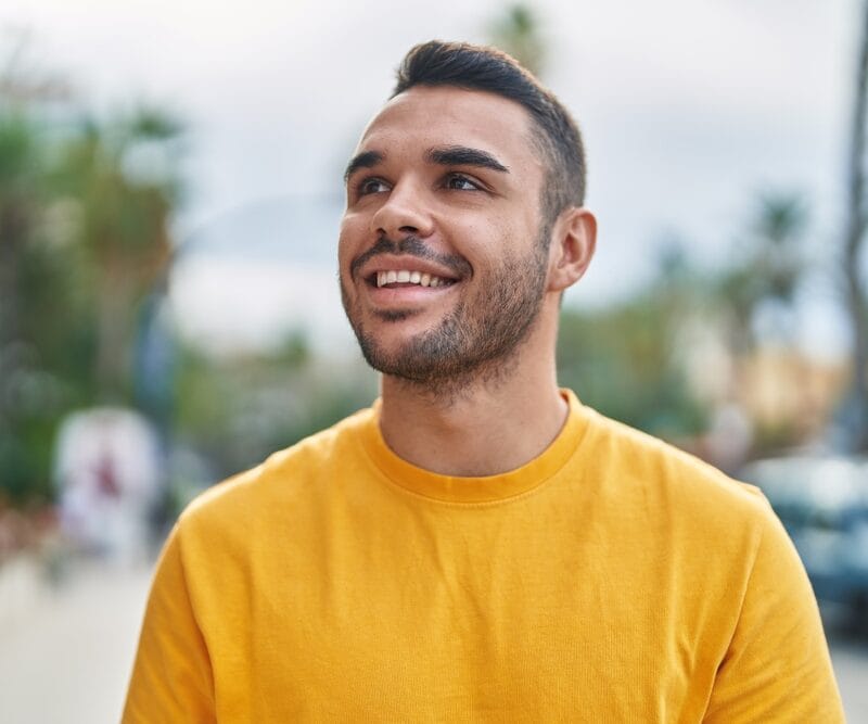 Young man wearing a bright yellow shirt smiling and looking up while walking along a palm tree-lined street with a calm, confident expression - how to stop dental implant bleeding