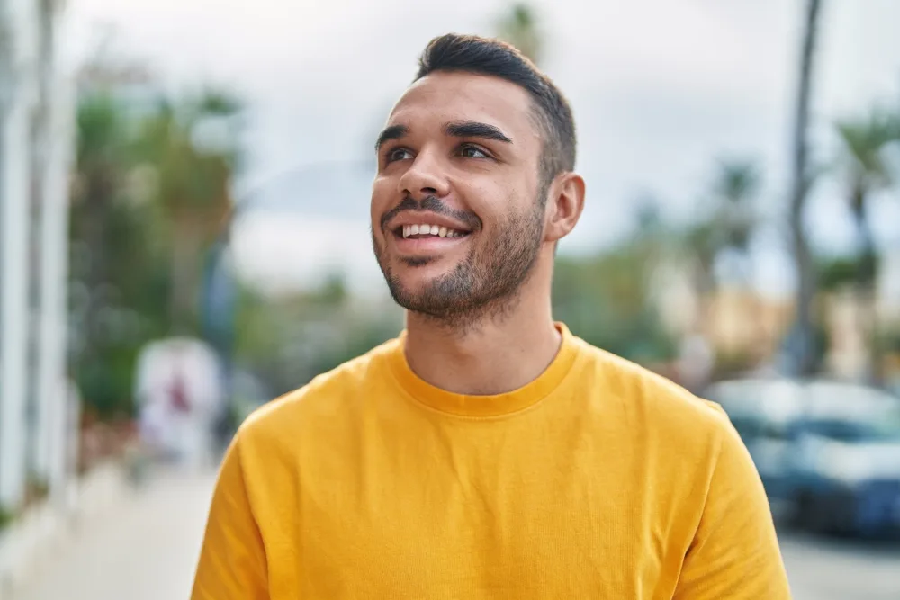 Young man wearing a bright yellow shirt smiling and looking up while walking along a palm tree-lined street with a calm, confident expression - how to stop dental implant bleeding