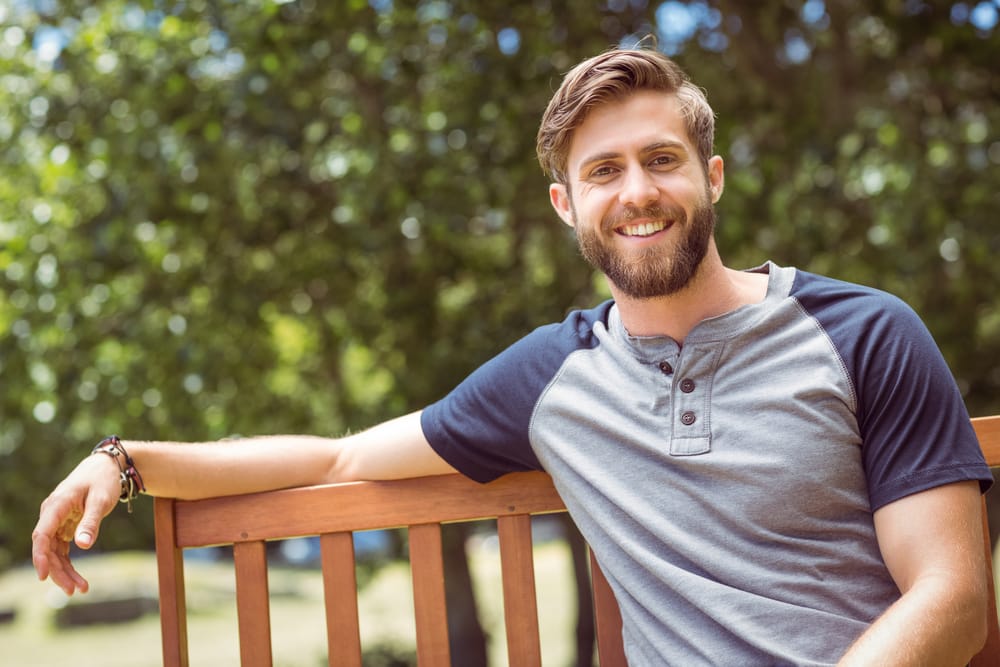 Young man with a beard and short hair wearing a casual henley shirt, smiling while sitting comfortably on a wooden bench in a sunlit park - Auburn Hills Family Dentistry