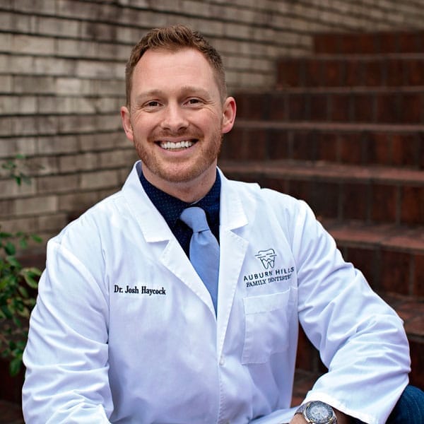 Dr. Josh Haycock, wearing a white coat and blue tie, smiling warmly while seated on outdoor steps with the Auburn Hills Family Dentistry logo visible on his jacket - Auburn Hills Family Dentistry
