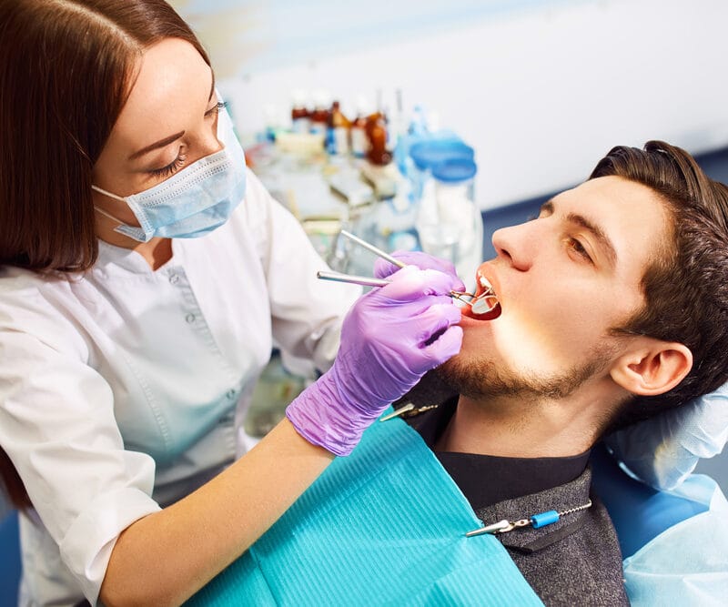 Female dentist wearing gloves and a face mask treating a male patient reclined in a dental chair, using tools during a dental procedure - how long does a root canal procedure take
