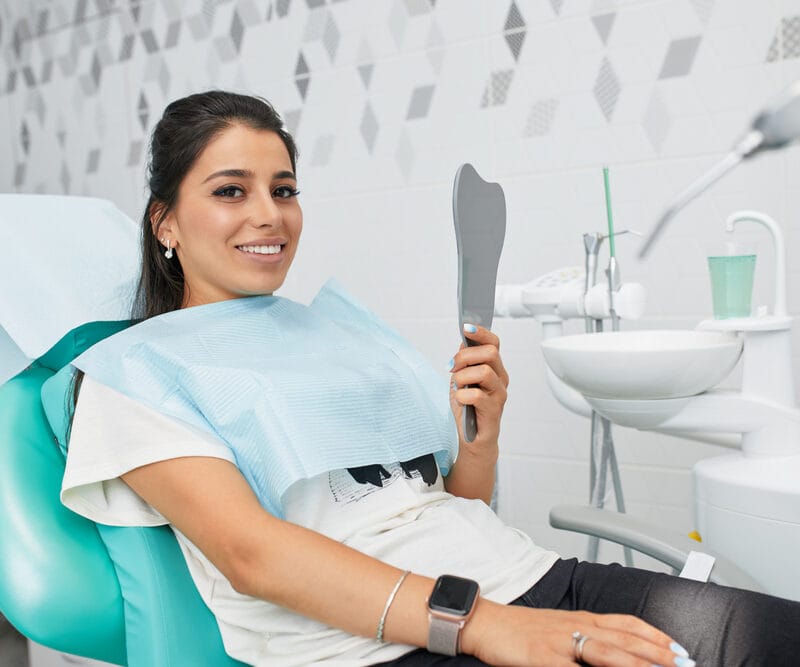 Young woman sitting in a modern dental chair with a bib on, smiling and holding a handheld mirror, appearing satisfied after a dental procedure - crown root canal