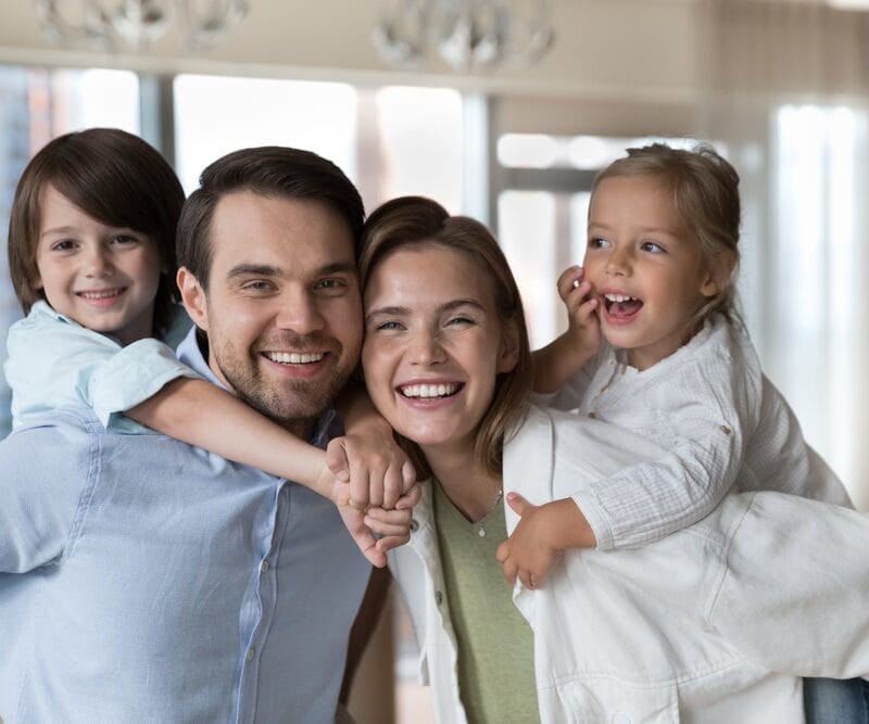 Happy mother and father standing close together indoors, each carrying a young child on their back, all smiling and enjoying playful family time - crown root canal