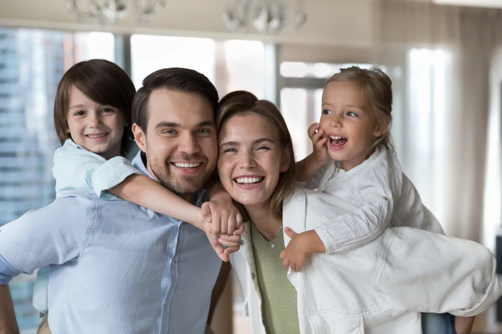 Happy parents standing close and smiling with their two young children playfully riding on their backs, enjoying time together in a modern, sunlit home - root canal going bad